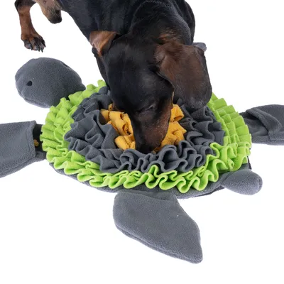 Hund spielt mit einer Schnüffelmatte in Schildkrötenform aus grauem, grünem und gelbem Stoff. Matte dient zur Beschäftigung und Leckerli-Suche.