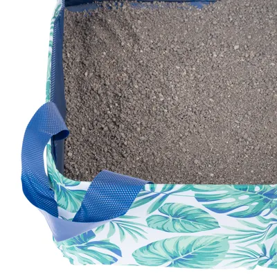 Przenośna torba z tropikalnym wzorem liści, wypełniona szarym żwirkiem dla kota. Widoczne niebieskie uchwyty ułatwiające transport.