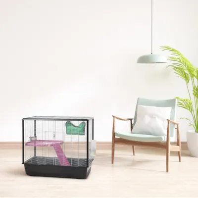 Cage pour petits animaux en métal et plastique noir, avec plateforme rose, maisonnette verte et abreuvoir, posée sur un sol en bois clair dans un intérieur moderne.