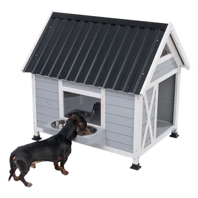 Gråt hundehus med sort tag, integreret foderområde og to skåle i rustfrit stål. En hund spiser af en skål foran huset.