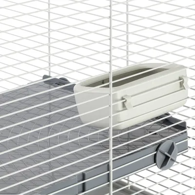 Gros plan sur une mangeoire en plastique gris clair fixée à l'intérieur d'une cage à barreaux blancs, posée sur un plateau en plastique gris.