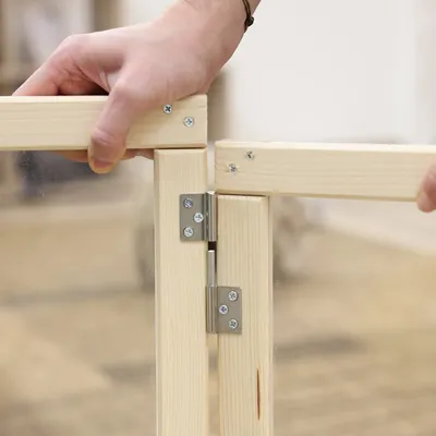 Montage eines Holzrahmens mit Scharnieren und Plexiglas. Hände halten die Teile zusammen, Schrauben sind sichtbar.