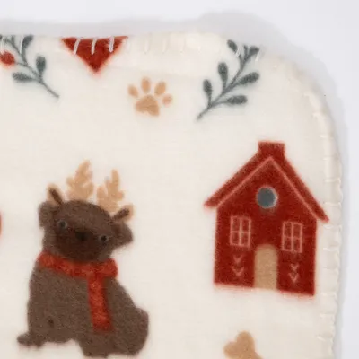 Dettaglio di una coperta color crema con la stampa di un cane con corna e sciarpa, una casa rossa, impronta di zampa e motivo vegetale. Dettaglio di una coperta color crema con la stampa di un cane con corna e sciarpa, una casa rossa, impronta di zampa e motivo vegetale.