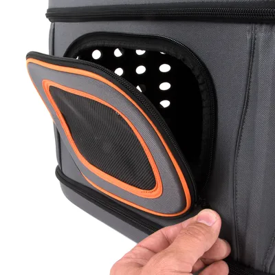 Sac de transport gris pour animaux avec ouverture zippée partiellement ouverte, filet noir et bordure orange, main tenant la fermeture éclair. Trous d’aération visibles à l’intérieur.