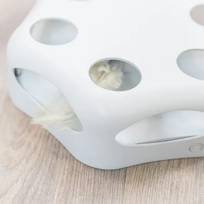 Bílý plastový předmět s otvory, uvnitř viditelné světlé peří. Umístěno na dřevěné podlaze.