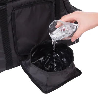 Main versant de l’eau d’un verre dans une gamelle pliante noire en tissu, posée devant un sac noir à poche zippée visible en arrière-plan.