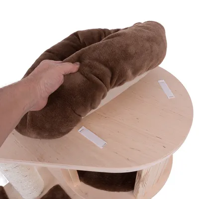 Hand plaatst bruin pluche kussen met klittenband op ronde houten plank van krabpaal. Kussen en bevestigingsstrips duidelijk zichtbaar.