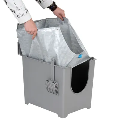 Kattenbak van grijs plastic met uitneembare zilverkleurige binnenzak en kattenbakschep aan de zijkant, hand pakt de binnenzak vast.