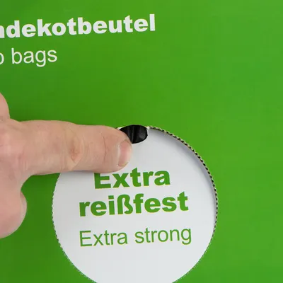 Tekst op verpakking: Extra reißfest, Extra strong. Hand pakt een zwarte hondenpoepzak uit de doos.