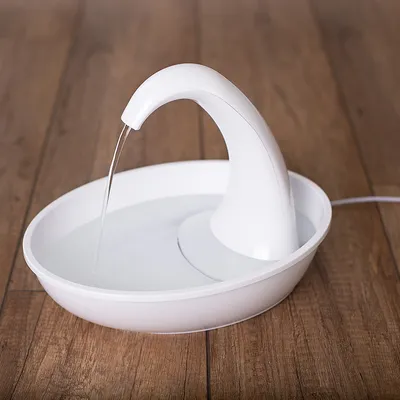 Fontaine à eau blanche en plastique avec bec courbé, jet d’eau continu dans un bol rond, posée sur un sol en bois. Câble d’alimentation visible à droite.