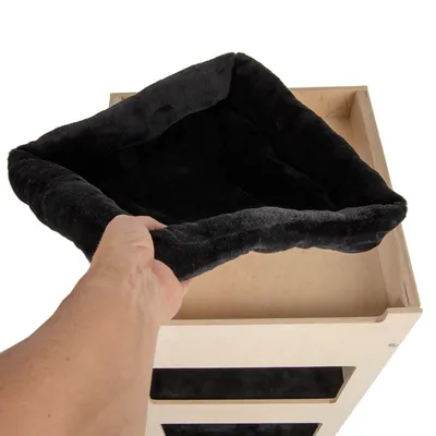 Ruka vkládá černý plyšový pelíšek do horní části dřevěného boxu. Viditelný materiál: textil a světlé dřevo.