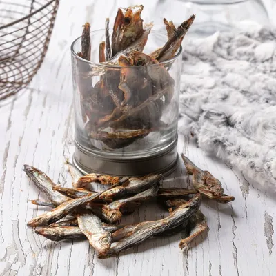 Dried whole fish treats displayed in a glass and scattered on a wooden surface, showing the product’s size and texture. Dried whole fish treats displayed in a glass and scattered on a wooden surface, showing the product’s size and texture.