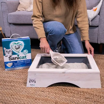 Sanicat Active White arena para gatos, sin olor. Mujer limpia el arenero con pala. El envase muestra detalles del producto y la imagen de un gato.