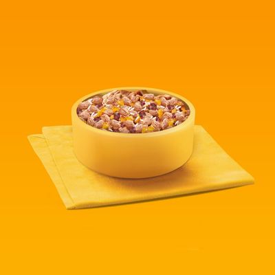 Cuenco amarillo con mezcla de pienso seco de trozos de carne y dados de verduras sobre paño amarillo, fondo naranja.