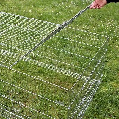 Metallgehege mit Gitterwänden, Deckel wird von Hand geöffnet. Gehege auf grünem Gras platziert.