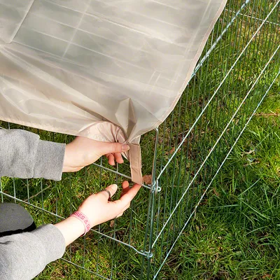 Montage d’une bâche beige sur un enclos extérieur en métal, mains attachant la bâche à la structure grillagée posée sur l’herbe.