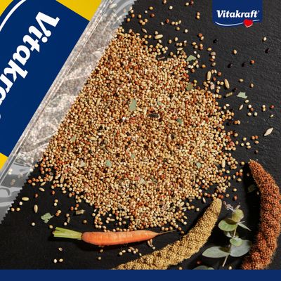 Mezcla de alimento Vitakraft con varios granos, una zanahoria, mijo y hojas. El logotipo de Vitakraft es visible en el envase.