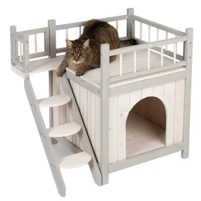 Casa de madera para gatos con escalera, balcón y zona de descanso. Gato tumbado en la plataforma superior. Casa de madera blanca y gris con entrada abierta.