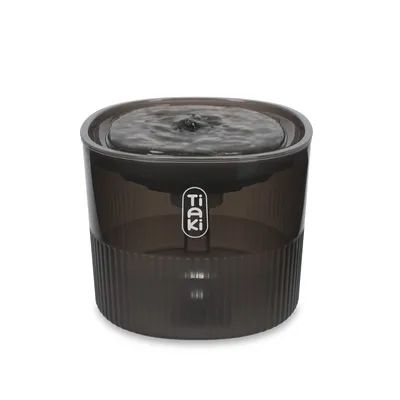 Fontaine à eau en plastique transparent fumé, marque Tiaki, avec écoulement d’eau visible sur le dessus. Fontaine à eau en plastique transparent fumé, marque Tiaki, avec écoulement d’eau visible sur le dessus.