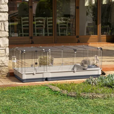 Grosser Käfig mit Metall-Gitterwänden und Kunststoffboden, ausgestattet mit Heuraufe und Trinkflasche. Geeignet für den Aussenbereich, abgebildet auf einer Terrasse mit Grasfläche.