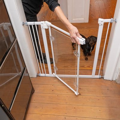 Biela bezpečnostná bariéra s dverovým otvorom, namontovaná v dvernom ráme. Osoba otvára dvere, za nimi stojí malý pes na drevenej podlahe.