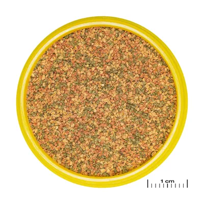 Detailní záběr na barevné granulované krmivo ve žluté nádobě, s měřítkem pro porovnání velikosti (1 cm).