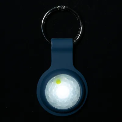 Porte-clés rond avec lumière LED blanche allumée, boîtier en plastique bleu foncé et anneau métallique visible en haut de l’image.