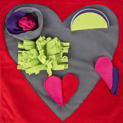 Snuffle mat in heart shape with grey and red fabric, colourful fleece elements such as green fringes, pink hearts and hideable treat pockets.