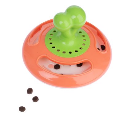 Jouet distributeur de croquettes en plastique orange et vert, forme ronde avec poignée en os, croquettes visibles à l’intérieur et quelques-unes dispersées autour.