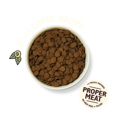 Bowl of dry pet food. Visible text: 'PROPER MEAT with freshly prepared, no meat meal or fillers'. Faint text: 'Much LOVED RECIPE'. Small olive illustration beside bowl.