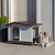 Caseta para perro de plástico gris y marrón con techo a dos aguas, puerta amplia y base elevada. Perro de tamaño mediano tumbado en la entrada de la caseta.