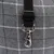 Close-up of a black strap with adjustable buckle and silver carabiner hook in front of grey fabric with check pattern.