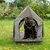 Bruine hond zit in een grijze, draagbare tent met metalen frame op een grasveld. Tent heeft een open deur en zijventilatie.