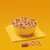 Bowl filled with mixed dry pet food pieces, including brown kibble, yellow chunks, white rice-shaped bits and dark cubes, shown on a yellow napkin with some pieces scattered in front.