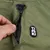 Poche zippée sur tissu vert avec badge Tiaki visible, main ouvrant la fermeture éclair. Cordon de serrage noir en haut de l’image.