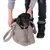 Petit chien noir et marron assis dans un sac de transport en tissu beige, tenu par une personne portant un pull gris et des bottes noires. Laisse grise attachée au collier du chien.