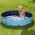 Piscina redonda azul para perros colocada sobre césped, con un perro de tamaño grande saltando dentro del agua. Material plástico visible y desagüe en la parte frontal.
