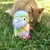 Plush toy in llama shape with colourful fur, sitting on a meadow. In the background, an orange cat in the grass.