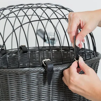 Cestino in vimini con griglia metallica nera e cinturini in pelle, mani che chiudono una fibbia sul bordo superiore.