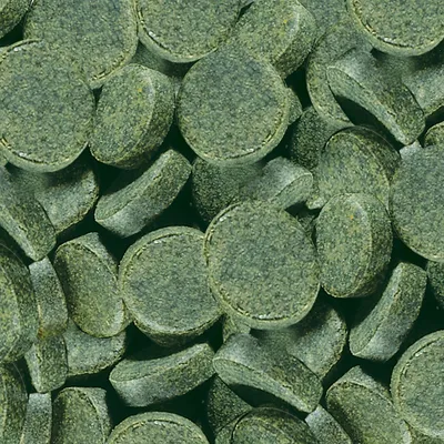 Tabletas verdes con superficie rugosa, posiblemente complemento alimenticio o aditivo para pienso. Forma redonda y tamaño compacto visible.