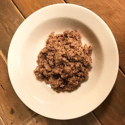 Annos ruskeaa, karkeaksi jauhettua lemmikin märkäruokaa valkoisella posliinilautasella puupöydällä. Tuotemerkki ja tarkempi tuotenimi eivät näy kuvassa.