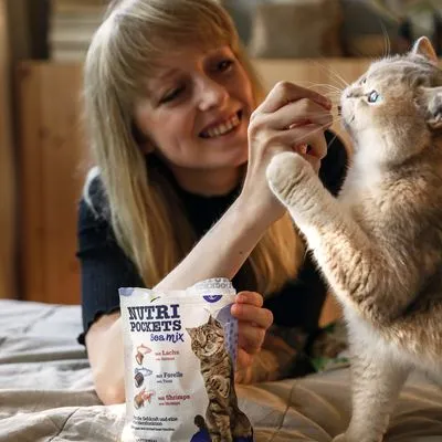 Kat krijgt snack uit zak Nutri Pockets sea mix; verpakking toont kat en tekst: met Lachs, Forelle, Shrimps. Persoon houdt de snack vast voor de kat.