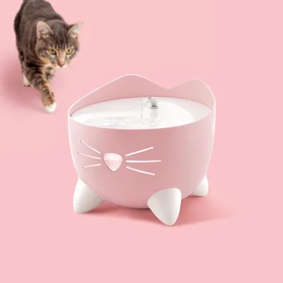 Rosa Trinkbrunnen in Katzenform mit fliessendem Wasser, dekorative Schnurrhaare und Pfoten. Katze im Hintergrund auf rosa Hintergrund.