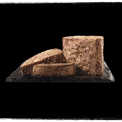 Sliced block of brown wet pet food on a dark stone surface, showing texture and cut sections. No visible packaging or branding.
