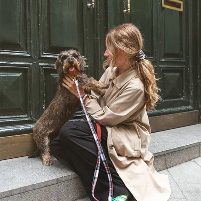 Vrouw met lang haar en beige jas zit op een stoep en houdt een kleine, ruwharige hond aan een rood-wit-blauwe riem vast voor een groene deur.