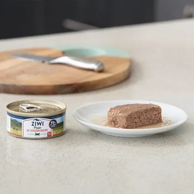 Boîte de pâtée pour animaux Ziwi Peak NZ Venison Recipe 92 %, à côté d'une portion servie sur une assiette blanche. Texte en anglais visible sur l'emballage.
