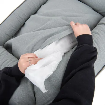 Mani che aprono la fodera di un cuscino o letto per animali in tessuto grigio, mostrando l'interno imbottito con materiale bianco visibile.