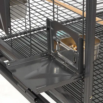 Sportello aperto in metallo di una gabbia con ciotola interna in acciaio inox contenente mangime, griglia e posatoio in legno visibili.