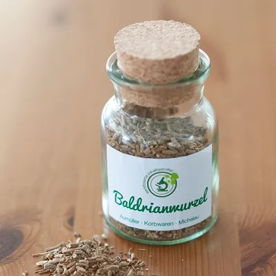 Glazen fles met kurksluiting, gevuld met valeriaanwortel. Etiket toont 'Baldrianwurzel', 'Aurmüller Korbwaren Michelau' en een groen logo met plant.