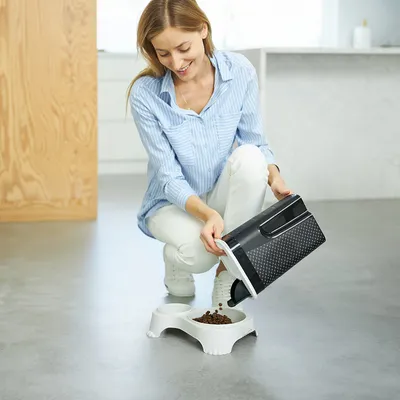 Person pouring dry pet food from a storage container into a double feeding bowl on the floor, demonstrating use of the container for dispensing food.
